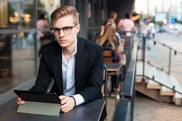 Young handsome blond guy sits with a tablet on the terrace of a cafe and looks at the camera