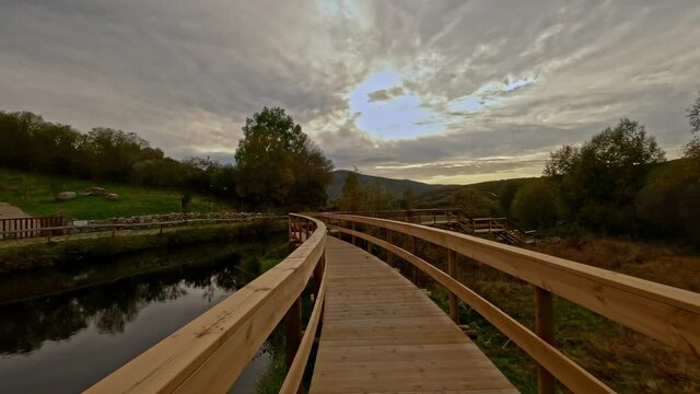 Moving forward from a wooden footbridge in Branda de Aveleda, Portugal, with the river and trees on a cloudy day, Parque Nacional da Peneda Geres, Portugal. At the end cow drinking in the river.