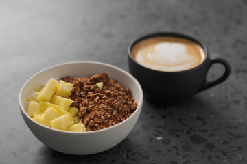 chocolate granola with mango and greek yogurt in white bowl with black cup with cappuccino on concrete background