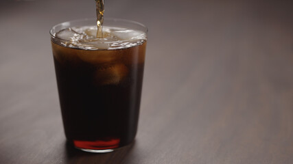 pour cola over ice cubes into tumbler glass on walnut table with copy space