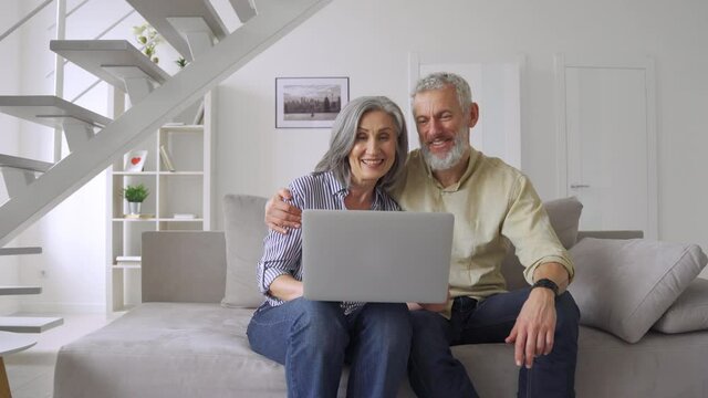 Happy middle aged mature couple waving greeting family having virtual meeting using laptop computer at home. Cheerful senior old adults grandparents talking, laughing, enjoying digital video call.
