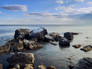 huge stones lie on the coast of the sea, the sea is calm, there are clouds in the sky, two cormorants are sitting on the rocks in the distance