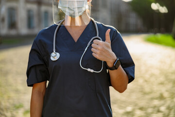 Naklejka premium Cropped shot of young woman nurse in uniform wearing face shield and mask for safety to prevent Covid19, showing thumbs up, standing outdoors on a sunny day
