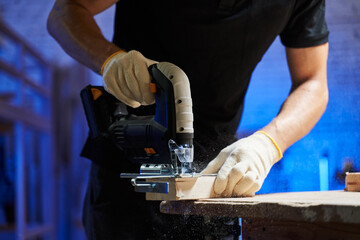 male hands in gloves on the table saw off a piece of board with a jigsaw - obrazy, fototapety, plakaty