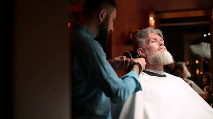 A gray-haired respectable man gets a beard trimming service in a barbershop. Men's personal care salon