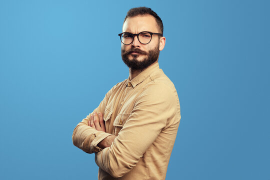 Image of serious young bearded guy looking at camera with crossed hands, isolated over blue background wearing eyeglasses and beige shirt