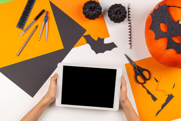 Preparing for Halloween. Teenage kid hands holding digital tablet computer. Colored paper, scissors, pumpkin, Halloween party decorations on white desk. Top view