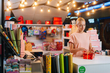 Medium shot of young sales woman holding and using golden ribbon to tie bow for red wrapped gift box for Christmas present at counter of holiday shop, blurred background of bright lighting.