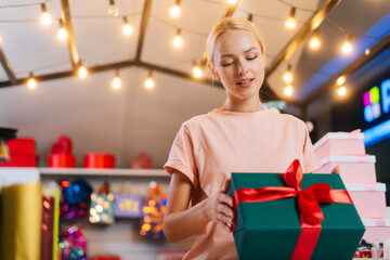 Low-angle view of young attractive saleswoman designer wrapping Christmas gift box tying red ribbon and decorating beautiful bow at counter of holiday store, blurred background of bright lighting.