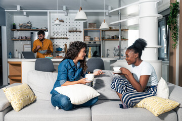 Two young woman sitting on the sofa drinking tea or coffee talking about female problems while man standing in the kitchen and cooking lunch for them reading recipe from the internet on laptop 