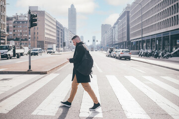 Young bearded caucasian man walking outdoor city crosswalk using smartphone wearing backpack, traveller checking map or commuter going work reading emails - obrazy, fototapety, plakaty