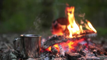 A metal mug with a drink stands next to the fire. Picnic in a camp in the woods. Fireplace in the camp, twilight