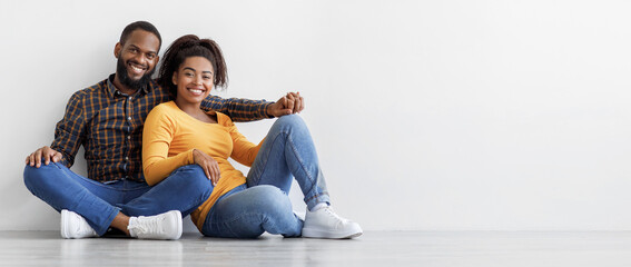 Smiling calm millennial african american couple, owners new apartment sit on floor on gray wall...