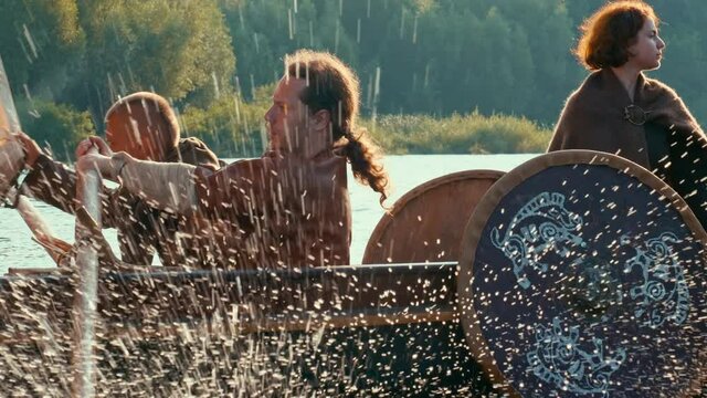 A Viking Man Stands Aboard the Drakkar Dressed in Armor and Looks into the Distance. Vikings Sail on an Old Ship with a Raised Sail Along a Calm River Against the Backdrop of a Rocky Coastline.