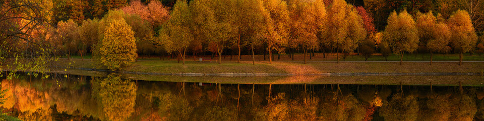 vibrant colors of october in beautiful autumn landscape. wide picturesque panoramic view of orange evening city park along the coast on the with lush trees reflected in the water