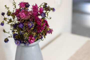 wilted flowers, chrysanthemum in different colors,purple, pink and blue, on a table in kitchen and on the window.beautiful light blue vase.home interior.flowers left,forgotten in vase,no water.end