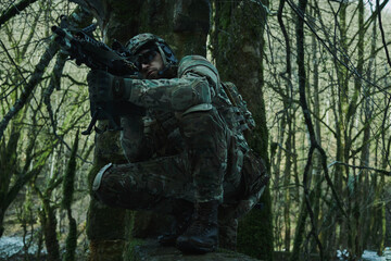 Fototapeta na wymiar Portrait of airsoft player in professional equipment in helmet aiming at victim with gun in the forest. Soldier with weapons at war