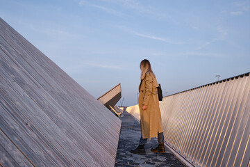 A girl in a trench coat stands on a modern bridge in the city. Tourist walks in Europe. Urban lifestyle. Travel and Adventure