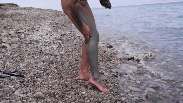 Young woman in beach of Dead Sea put curative mud on legs. Vacation, treatment and recreation in israel on Dead Sea. Salt and mud are good for health.