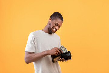 Money saving concept. Happy african american man putting banknotes into wallet and smiling, yellow...