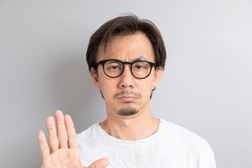 Adult Asian man with eyeglasses raising hand with disagreement on grey background.