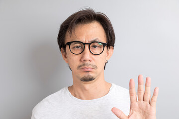 Adult Asian man with eyeglasses raising hand with disagreement on grey background.