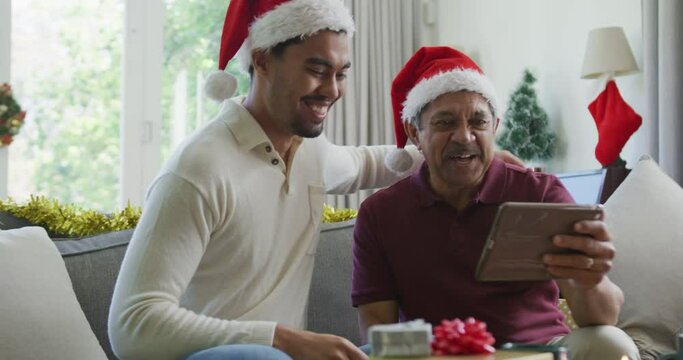Cheerful biracial son with senior father in santa hats making video call on tablet pc in christmas