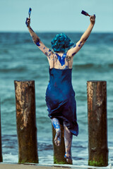 Happy woman performance artist in blue dress smeared with blue gouache paints dancing on beach
