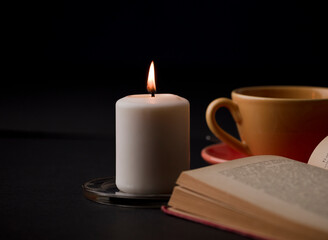 A candle lit with a flame, with a cup of tea with smoke, and a reading book in the background. (focus on candle).