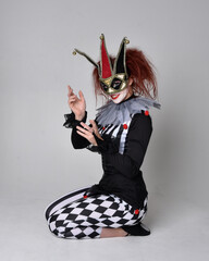 Full length  portrait of red haired  girl wearing a black and white clown jester costume, theatrical circus character.  Sitting down on floor, isolated on  studio background.