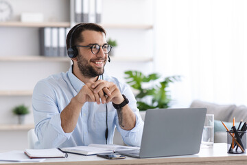Glad adult european male with beard in glasses, headphones works at laptop talk with clients in office or room interior