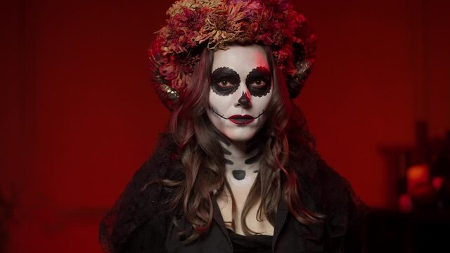 A brunette woman in a beautiful Calavera-style image on All Saints' Day performs a ritual in a dark room with red light from a campfire. Halloween makeup with ram horns, flowers, sewn mouth