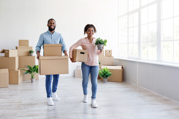 Fototapeta na wymiar Satisfied happy millennial african american male and female carry cardboard boxes with belongings in empty room
