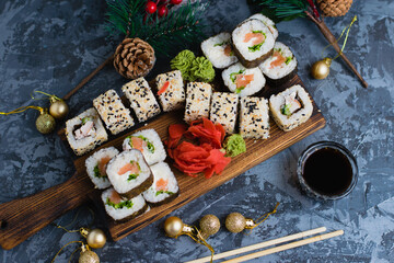 Large set of sushi on a wooden board top view. Rolls with sesame seeds, nori, cheese, red fish, cucumber and eel, ginger, wasabi, soy sauce, bamboo sticks and christmas decor on gray background