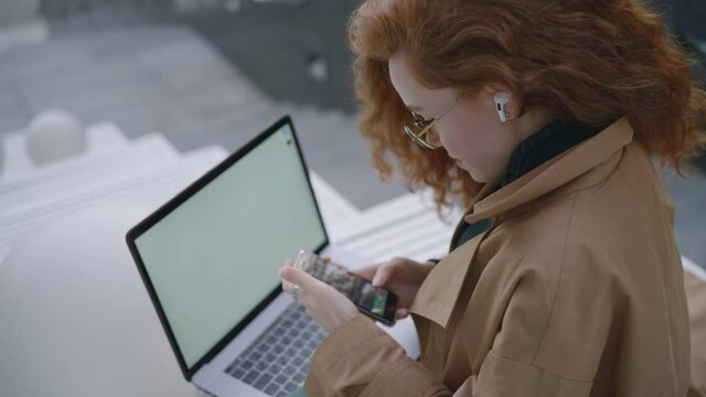 Office worker young woman in glasses puts on wireless bluetooth headphones in ears and starts to listen to music sitting on stairs ground with open laptop