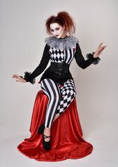 Fototapeta premium Full length portrait of red haired girl wearing a black and white clown jester costume, theatrical circus character. Sitting down on chair, isolated on studio background.