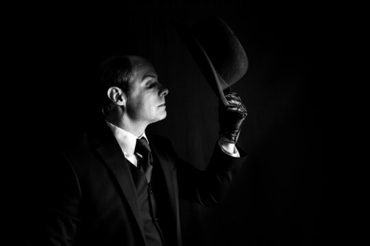 Profile Portrait of Butler in Dark Suit Doffing Bowler Hat on Black Background. Classic and Mysterious English Gentleman.