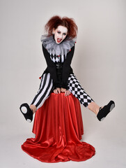 Obraz premium Full length portrait of red haired girl wearing a black and white clown jester costume, theatrical circus character. Sitting down on chair, isolated on studio background.