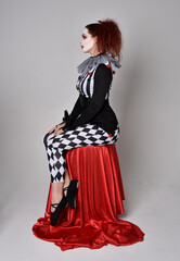 Full length  portrait of red haired  girl wearing a black and white clown jester costume, theatrical circus character.  Sitting down on chair, isolated on  studio background.