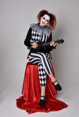 Obraz premium Full length portrait of red haired girl wearing a black and white clown jester costume, theatrical circus character. Sitting down on chair, isolated on studio background.