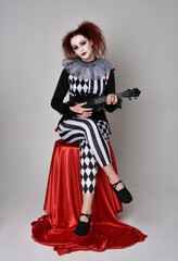Obraz premium Full length portrait of red haired girl wearing a black and white clown jester costume, theatrical circus character. Sitting down on chair, isolated on studio background.