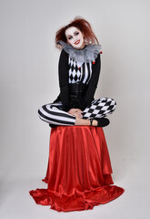Fototapeta premium Full length portrait of red haired girl wearing a black and white clown jester costume, theatrical circus character. Sitting down on chair, isolated on studio background.