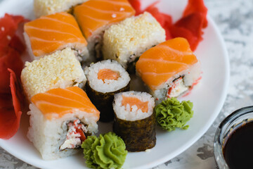 Sushi on a white plate on a light background with copy space. Delicious rolls with cheese, fish, sesame seeds, crab stick, wasabi, ginger and soy sauce.