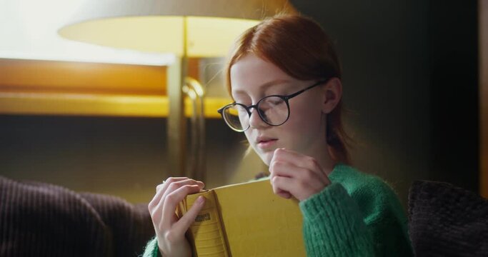 A girl in glasses reading a book with a dreamy look, sitting on the sofa