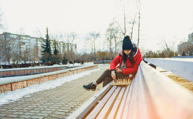 Young woman in winter clothes plays chess, sitting on bench in city park. Female in black cap with...