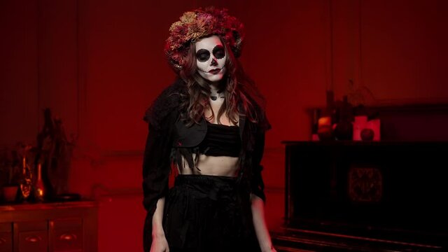 A woman in the form of a witch on a halloween with the horns of a ram and flowers on her head, bird's paws in her hands dancing in a dark room by the fire. Calavera Makeup for All Saints' Day