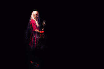 Scary young woman holding a candelabrum or candlestick, wearing a witch costume for Halloween, on black background - obrazy, fototapety, plakaty