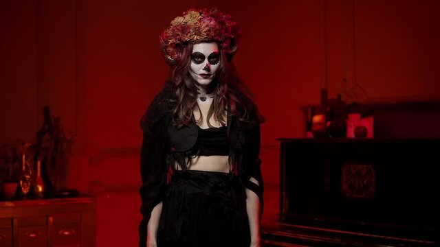 Atmospheric female image on a halloween in the style of the Spanish Day of the Dead. A brunette woman in witch rags, with ram horns, painted skull makeup in a dark burning room
