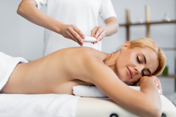 Obraz na płótnie Canvas masseur placing hot stone on blonde client in spa center