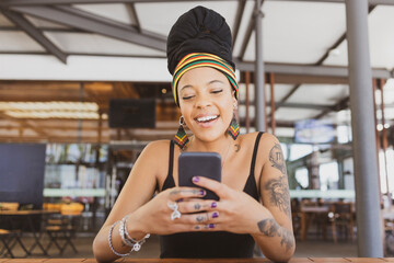 Beautiful tattooed African American woman wearing an african turban sitting at the table texting on her smartphone and smiling joyful. Copy space for text.